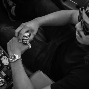 Top-down view of a man wearing a black beanie and sunglasses sitting in a brown leather car interior, holding casino poker chips near the gear shift.