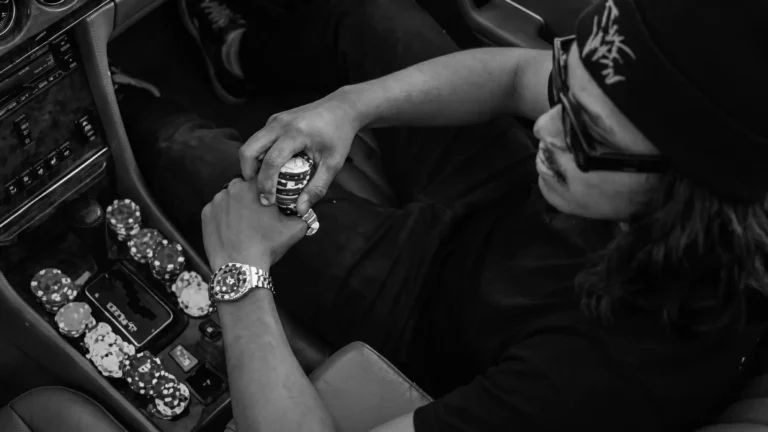 Top-down view of a man wearing a black beanie and sunglasses sitting in a brown leather car interior, holding casino poker chips near the gear shift.