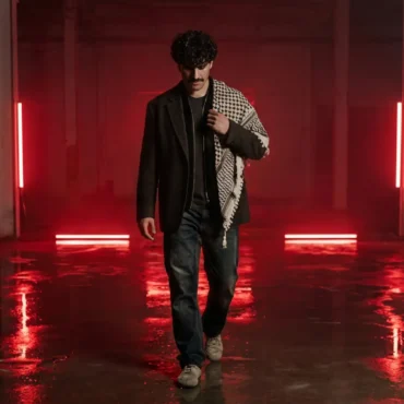 Dr Koul walking in a red neon lit warehouse wearing a keffiyeh.