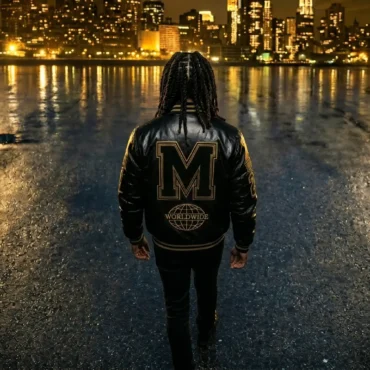 M'siyah from behind wearing a black leather varsity jacket facing a lit New York City skyline at night across the water.
