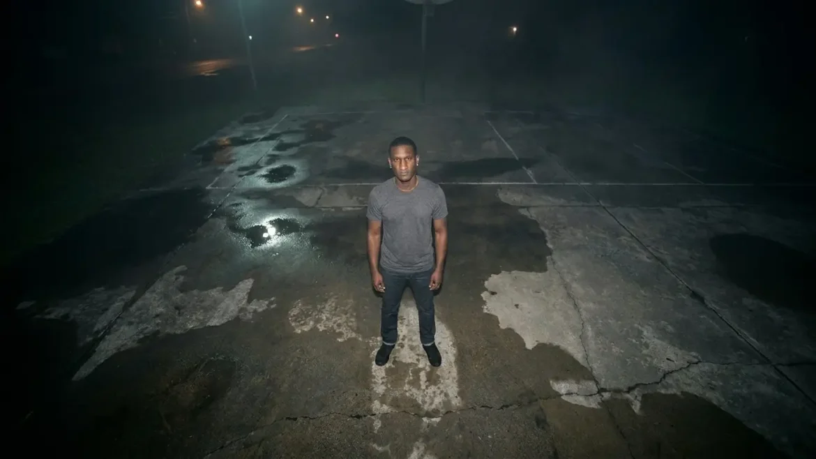 Hip hop artist T. Scales standing in the center of an outdoor concrete basketball court at night under dim streetlights.