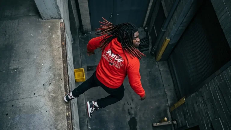 High angle view of rapper Wyn Calico in a red hoodie and black pants jumping across an urban concrete alleyway.