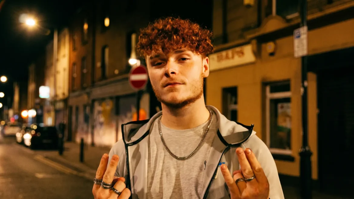 AC23 standing on a nighttime Newcastle street, curly red hair, grey hoodie over a t-shirt, layered silver chain, both hands raised in a ring-covered gesture toward the camera, promo image for Prince of the North