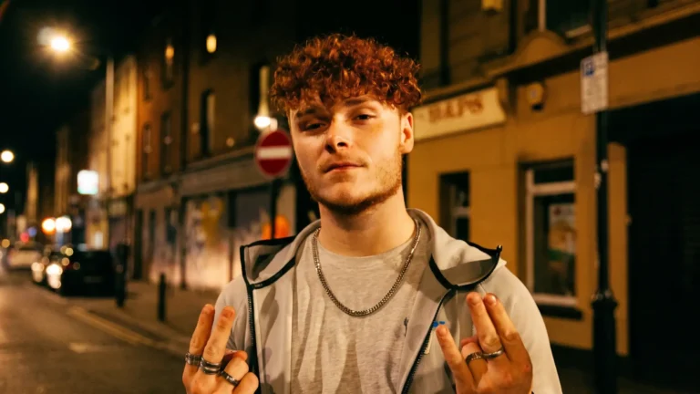 AC23 standing on a nighttime Newcastle street, curly red hair, grey hoodie over a t-shirt, layered silver chain, both hands raised in a ring-covered gesture toward the camera, promo image for Prince of the North