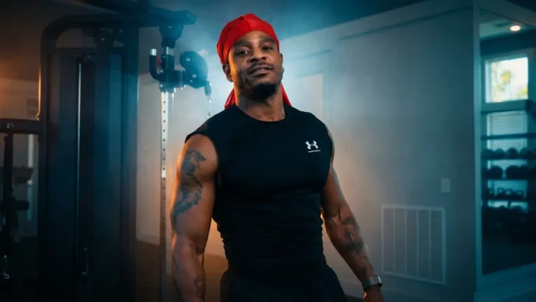 DaButta in red bandana and black tank top standing in a home gym, arms tattooed, looking directly at camera under blue-toned lighting