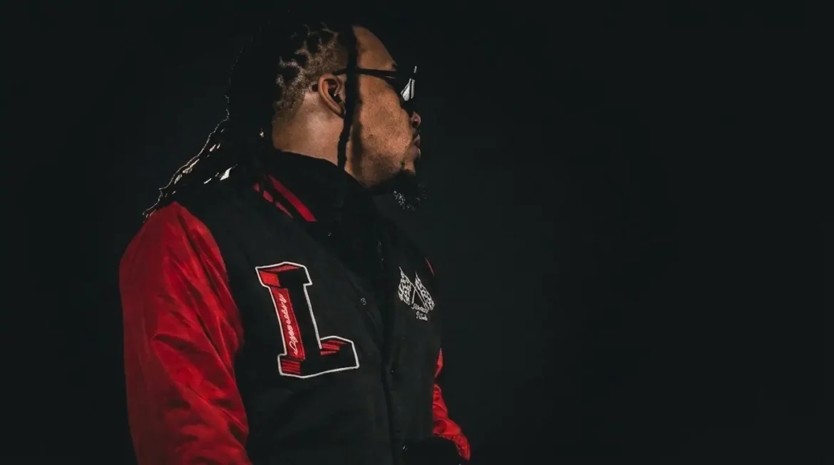 IanDakt in profile wearing a black and red varsity jacket, sunglasses on, standing against a dark background