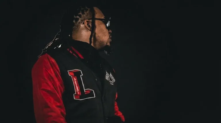 IanDakt in profile wearing a black and red varsity jacket, sunglasses on, standing against a dark background