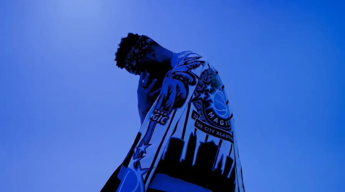 Low-angle shot of niceymost in a blue-toned Orlando Magic graphic jacket, head bowed, hand raised near his face, promoting his single "OKx2" off "4EVERDAT"