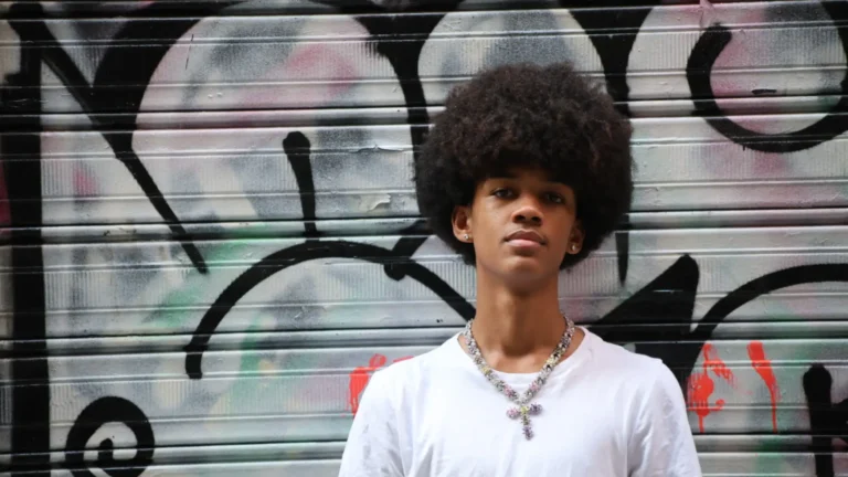 COLO, a young man with a large afro and a white t-shirt, wearing a pastel jeweled cross chain, standing in front of a graffiti-covered metal shutter, artist for "Radar"