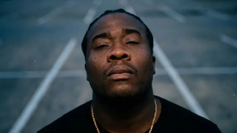Close-up portrait of Forward Next, artist behind "No Entities", chin raised with eyes half closed, wearing a black t-shirt and gold chain, standing on a parking lot with white painted lines at blue hour