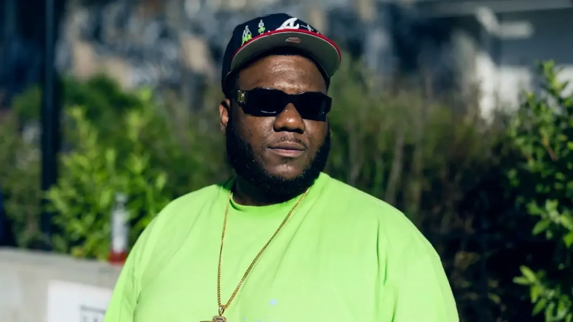 Close-up front-facing shot of Henry Hennessy, chest up, wearing a black fitted cap, dark sunglasses, and a bright green t-shirt with a gold chain, standing outdoors in front of green bushes