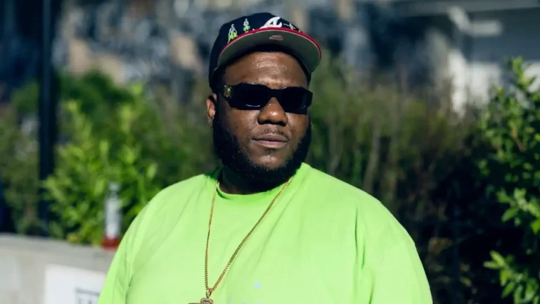 Close-up front-facing shot of Henry Hennessy, chest up, wearing a black fitted cap, dark sunglasses, and a bright green t-shirt with a gold chain, standing outdoors in front of green bushes
