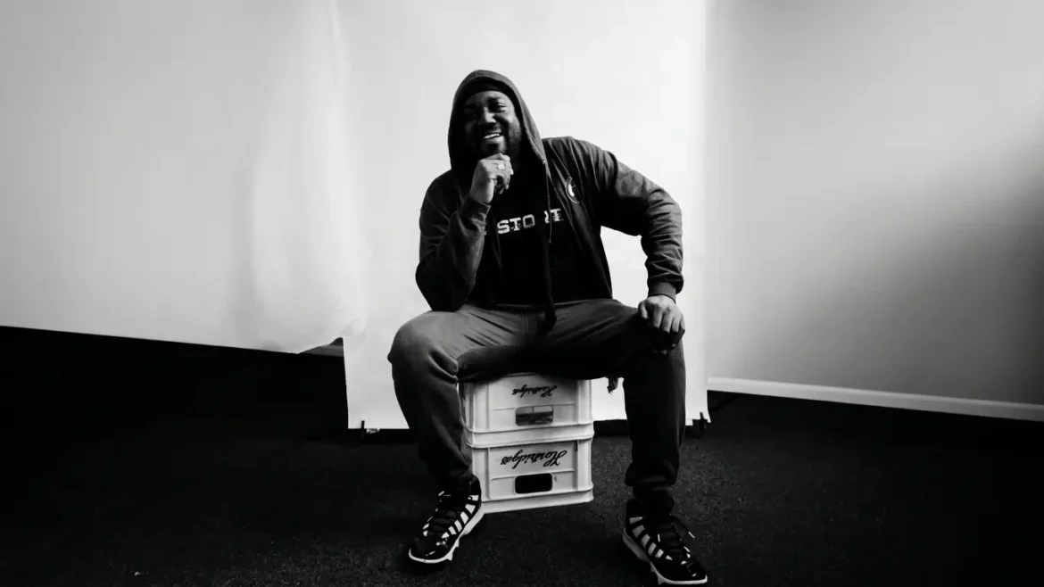 Black and white photo of Karnage The Great crouched on stacked crates, hood up, one hand resting under his chin, smiling