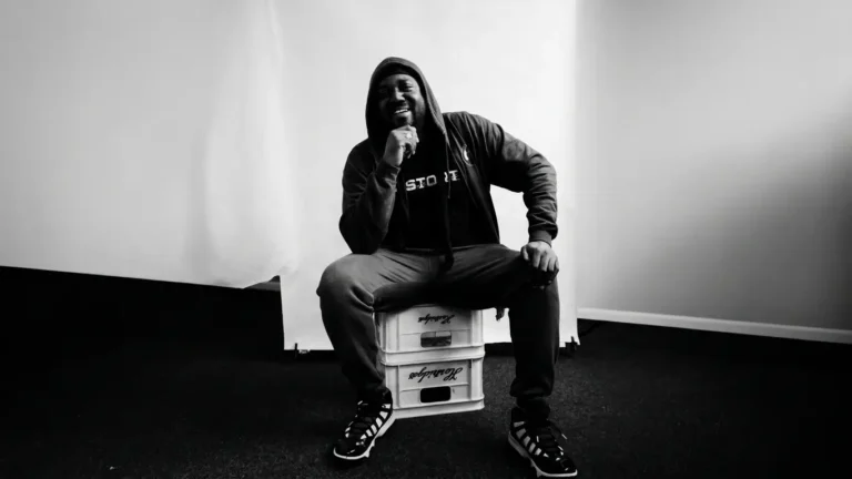 Black and white photo of Karnage The Great crouched on stacked crates, hood up, one hand resting under his chin, smiling
