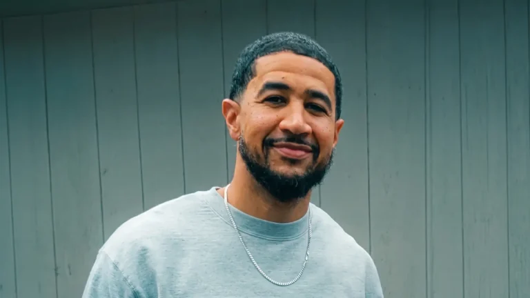 Leezy smiling at the camera in a grey sweatshirt and silver chain, standing against a grey wood-panel wall, promo photo for Dope On Purpose