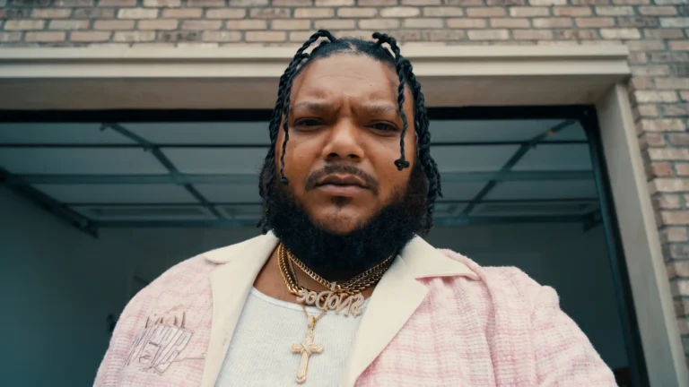 $No Doe, low-angle portrait in front of a brick building with glass garage doors, gold chains and cross pendant, pink tweed jacket, Bossa Nova