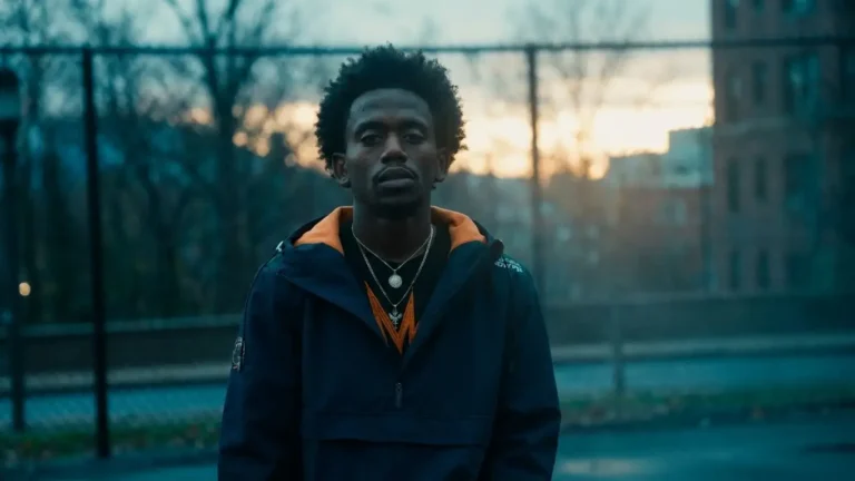 Wata standing outdoors at dusk in a navy and orange windbreaker, chain-link fence and city buildings behind him, direct gaze at camera, for "Queen's Cadence"