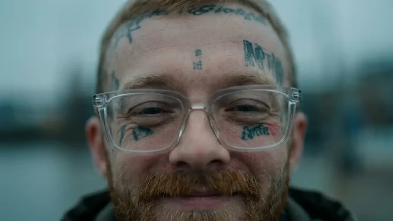 Close-up of Free Zoey smiling in clear-framed glasses, with face tattoos and a ginger beard, against a blurred grey-blue harbor, for the track “PROGRAM”