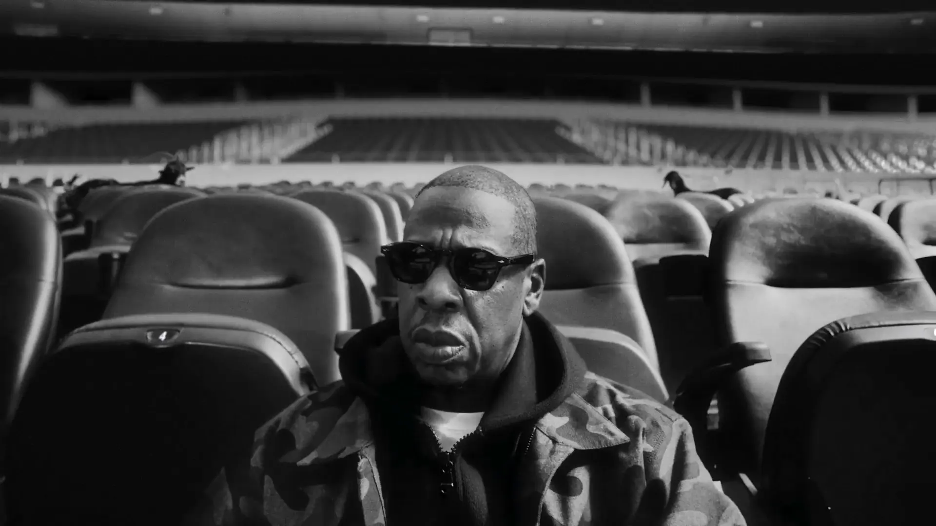 Jay-Z in sunglasses and a camo jacket sitting in empty stadium seats in the Super Bowl LXI halftime show announcement