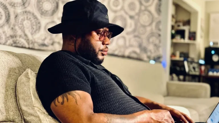 ShuPreme7 in side profile on a couch, wearing a black bucket hat, tinted glasses and a black knit polo, looking down at a laptop, for the track "She My Type"
