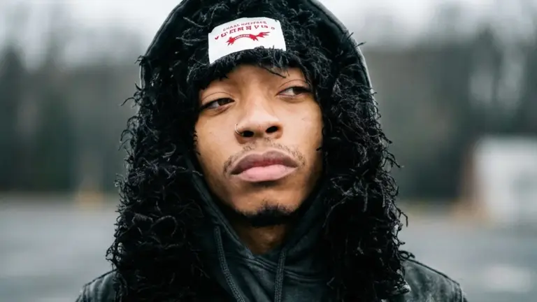 SPACEDOUTMARS in a black fringed hood with a white patch on the forehead and a nose ring, looking off to the side, blurred parking lot behind him, for the track “YA SHITE”