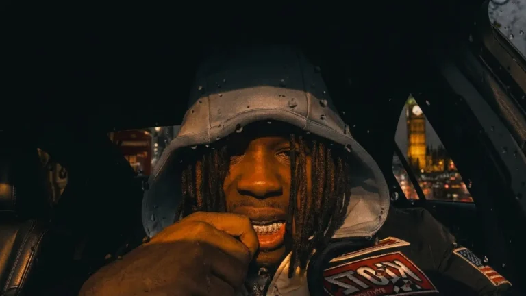 Trillz Cleanrun sits in a car at night with a rain-speckled grey hood up, locs falling over his forehead, one fist at his chin and a gold grill showing. The lit Elizabeth Tower and a red telephone box show through the windows behind him, track WHAT